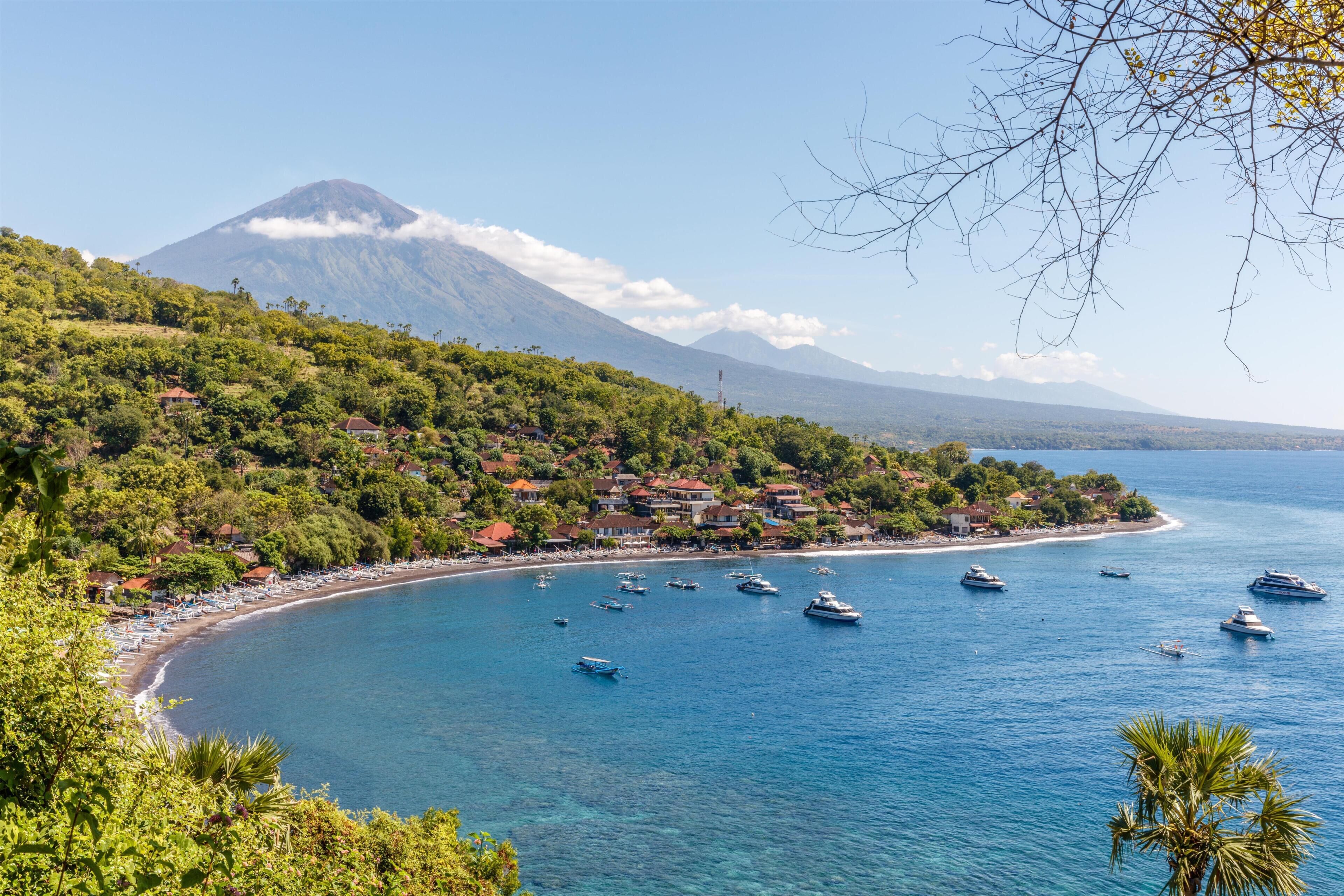 Un pueblo costero con una montaña al fondo, barcos en el agua y una playa con sombrillas.
