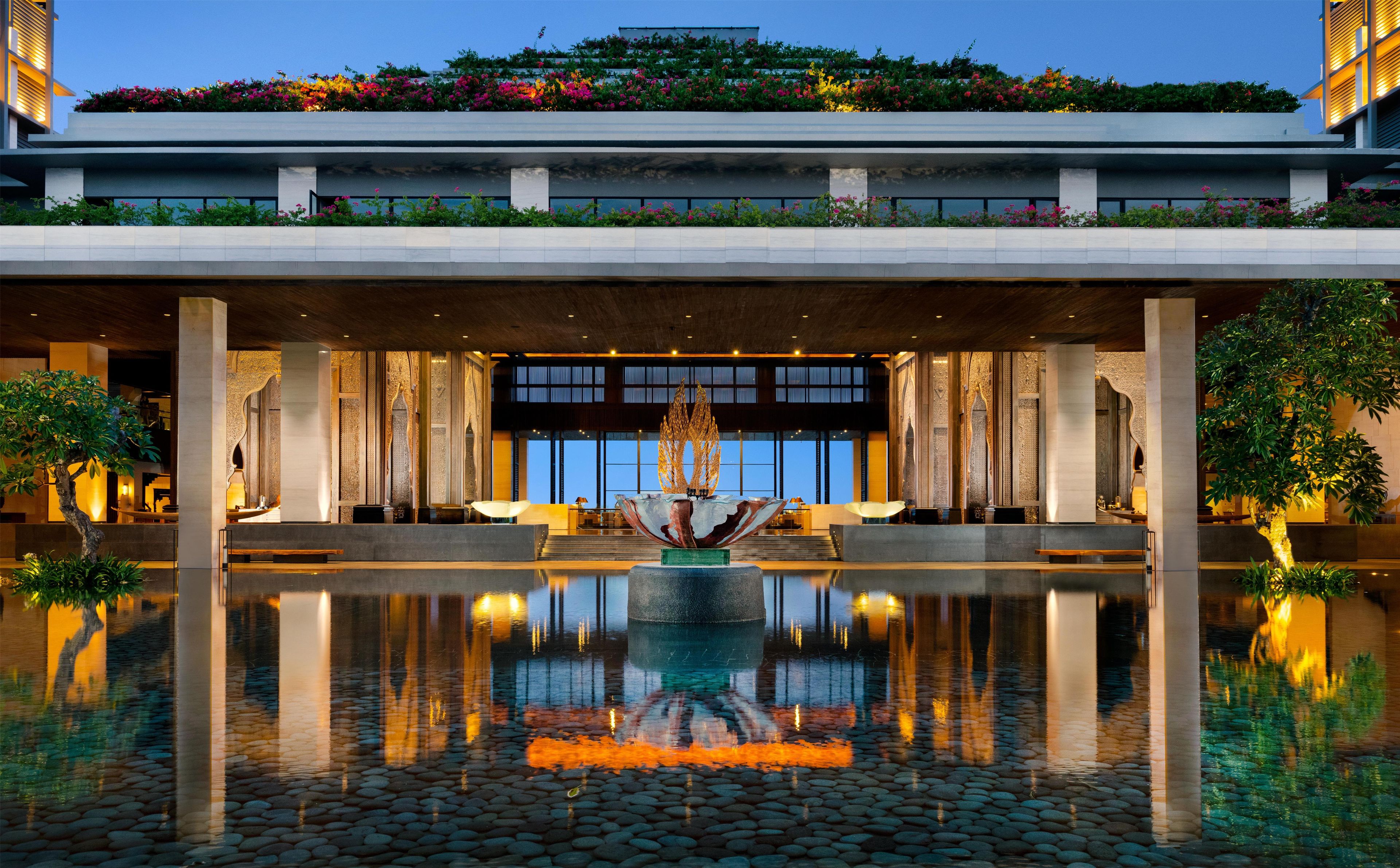 A modern hotel entrance with a reflective water feature, a seating area, and a large windowed facade.