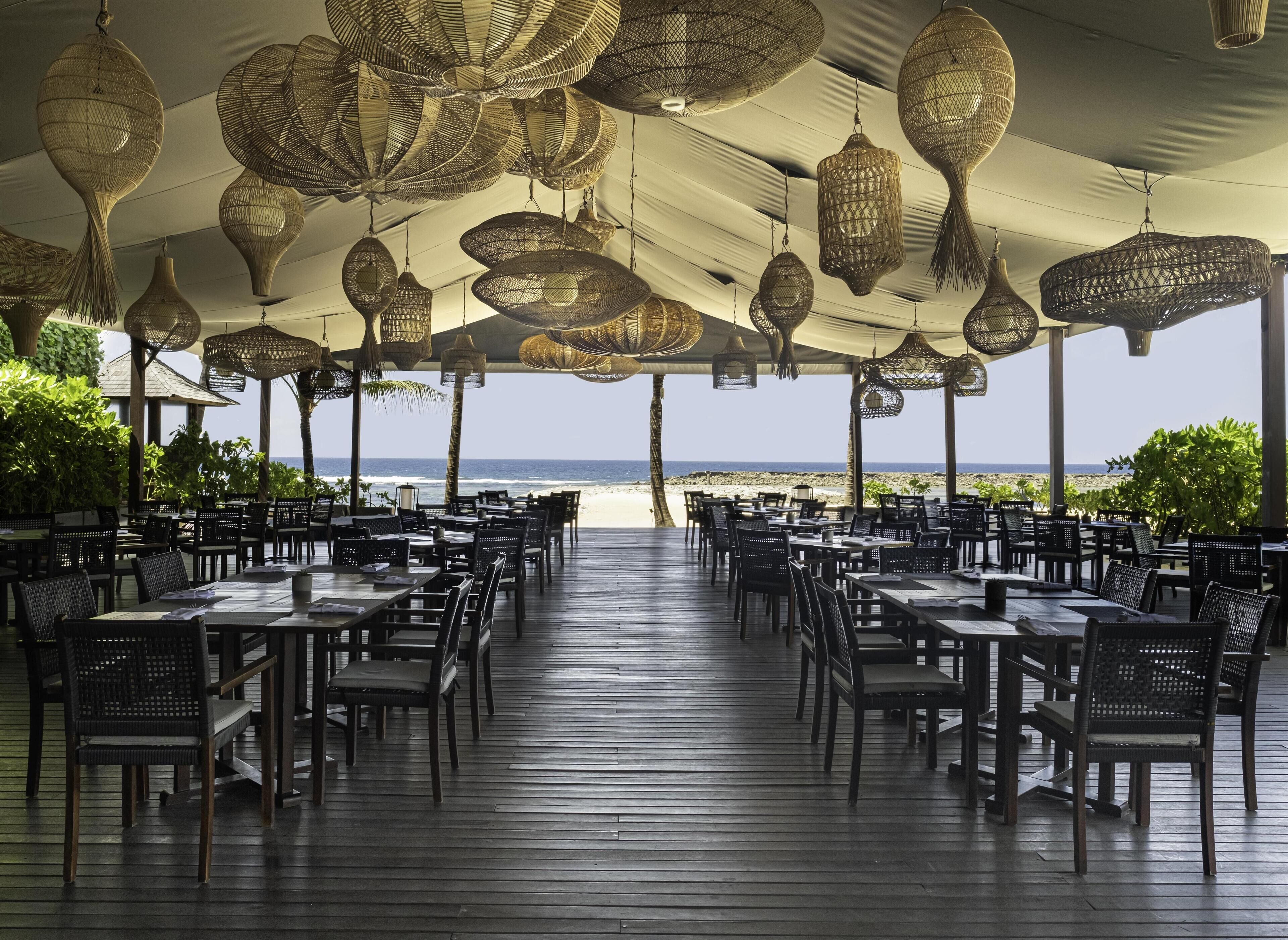 An outdoor dining area with tables and chairs arranged under a canopy, overlooking a beach.
