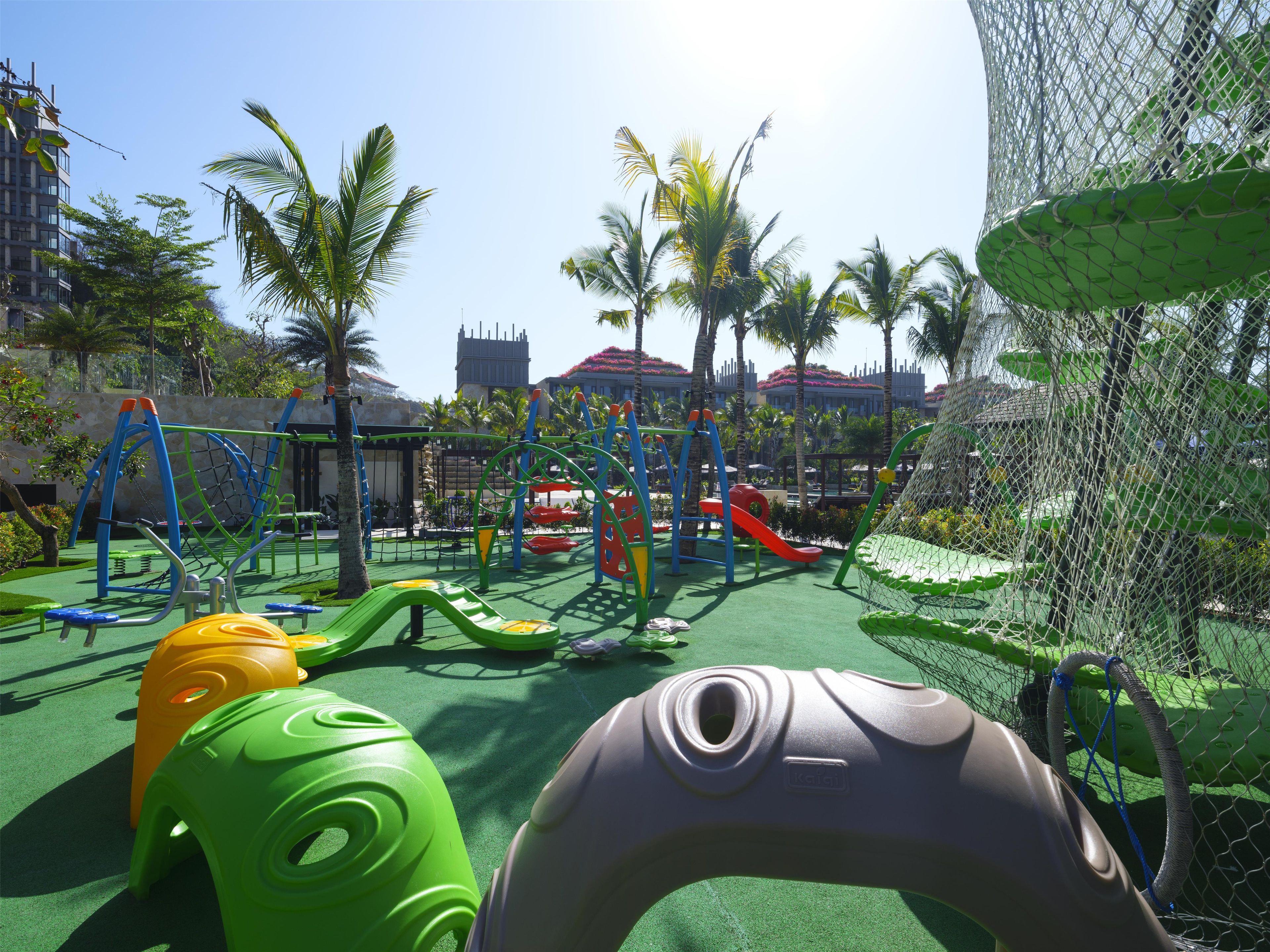 A playground with various play structures, surrounded by palm trees and a netted enclosure.