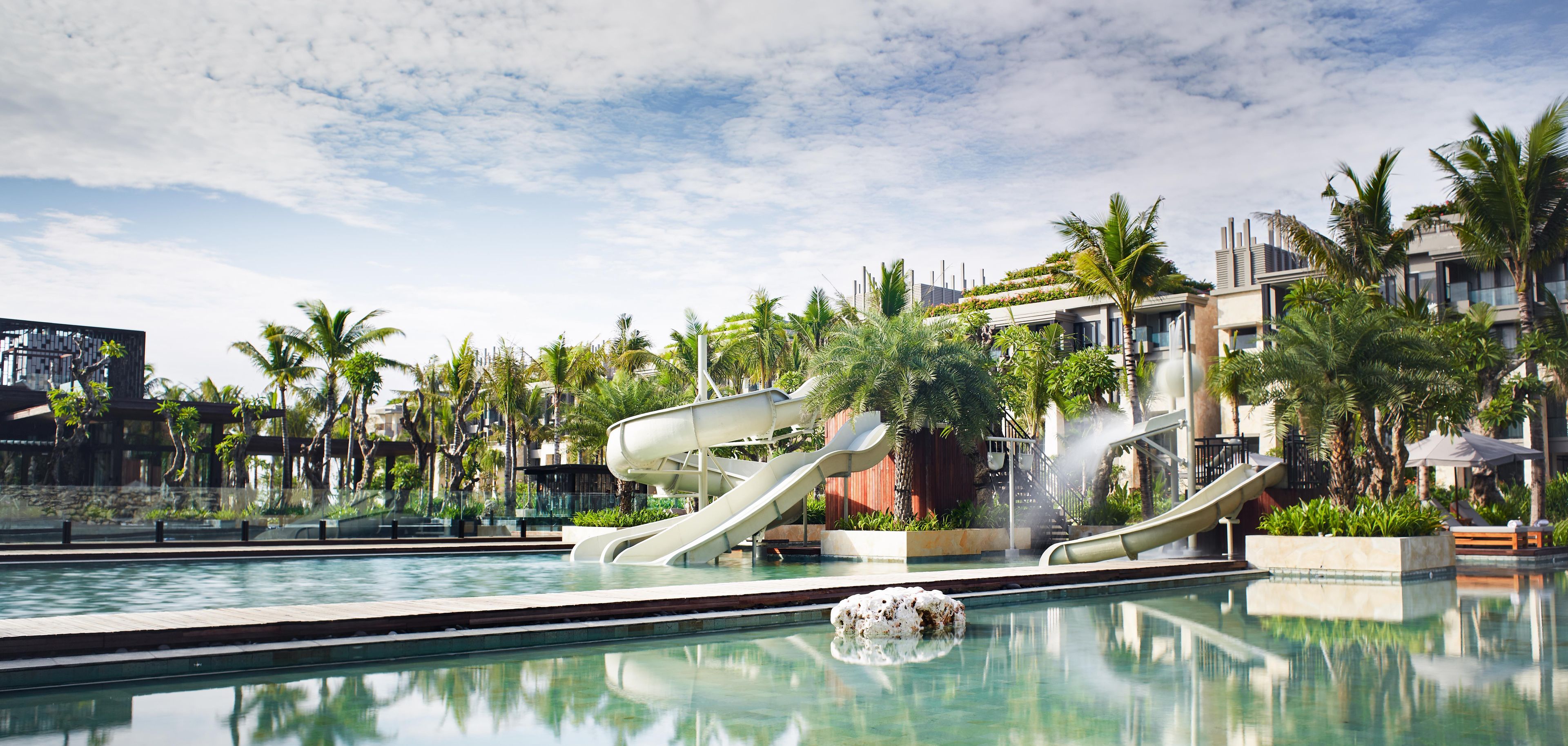 A swimming pool with multiple slides, surrounded by palm trees and modern buildings.