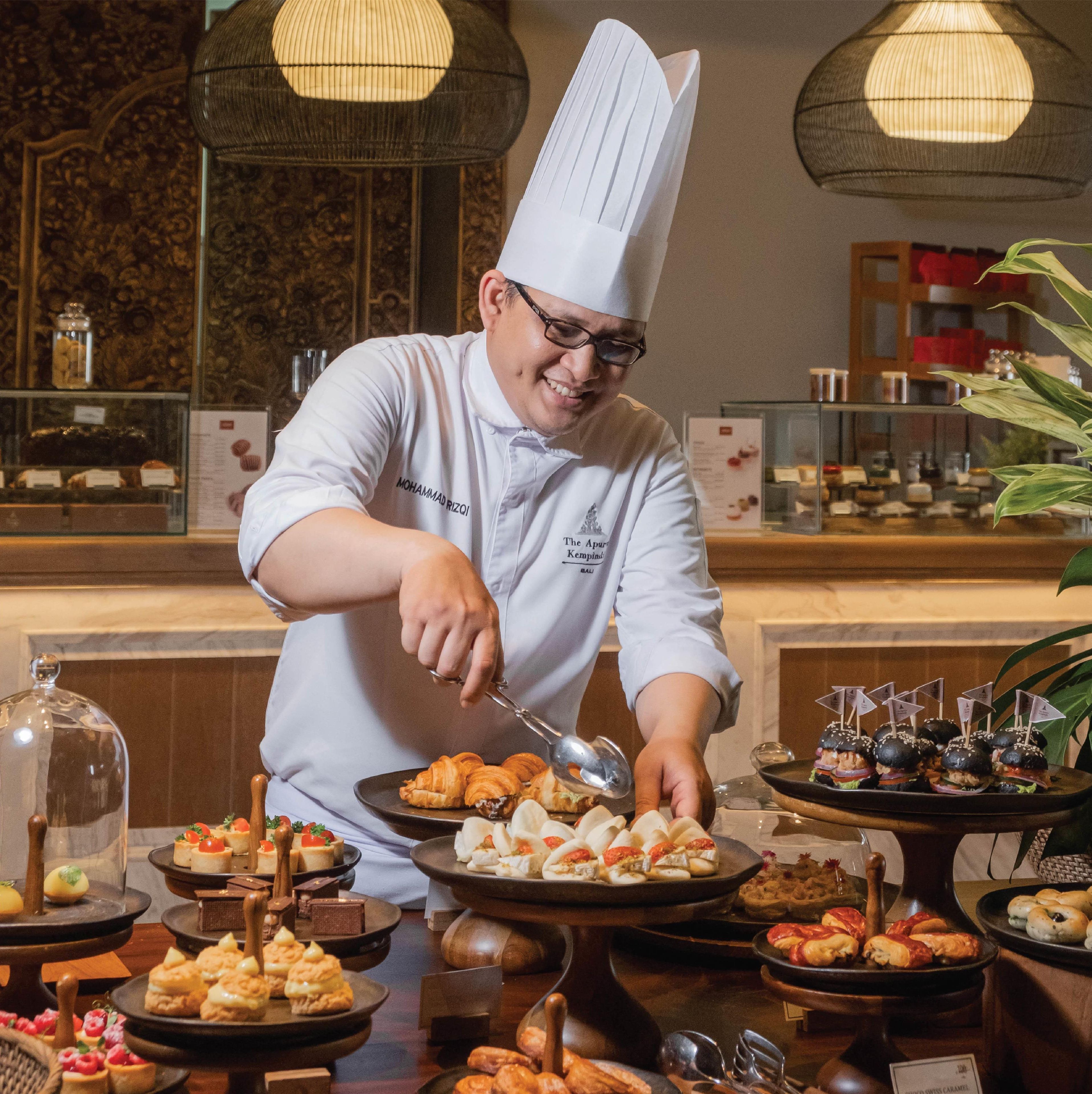 A chef in a white uniform with a tall hat, serving food at a buffet with various dishes displayed on plates.
