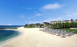 A beach resort with a sandy shore, lounge chairs, umbrellas, and a building with a modern design. - The Apurva Kempinski Bali (Nusa Dua)