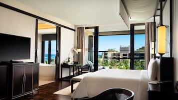 A modern hotel room with a large bed, a flat-screen TV, a desk with a chair, and a view of the ocean through large windows.