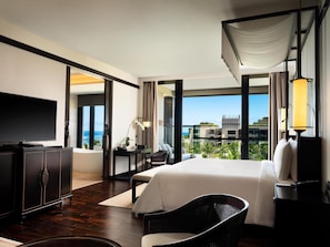 A modern hotel room with a large bed, a flat-screen TV, a desk with a chair, and a view of the ocean through large windows.