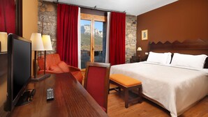 A hotel room with a bed, a desk with a TV and remote, a chair, and a view of mountains through a window.