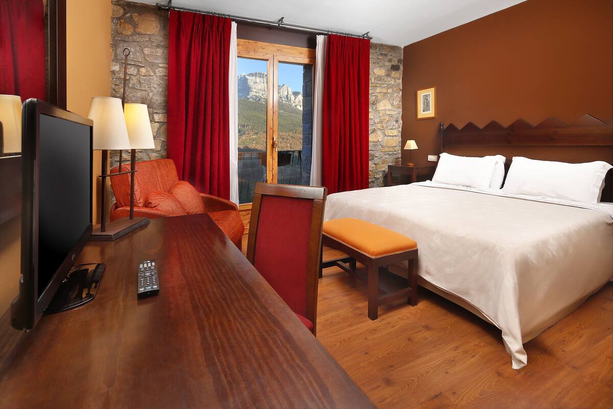 A hotel room with a bed, a desk with a TV and remote, a chair, and a view of mountains through a window.