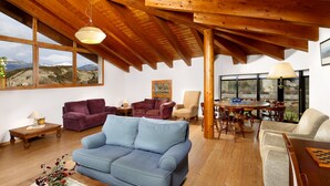A spacious living room with a blue sofa, wooden ceiling beams, and large windows offering mountain views.