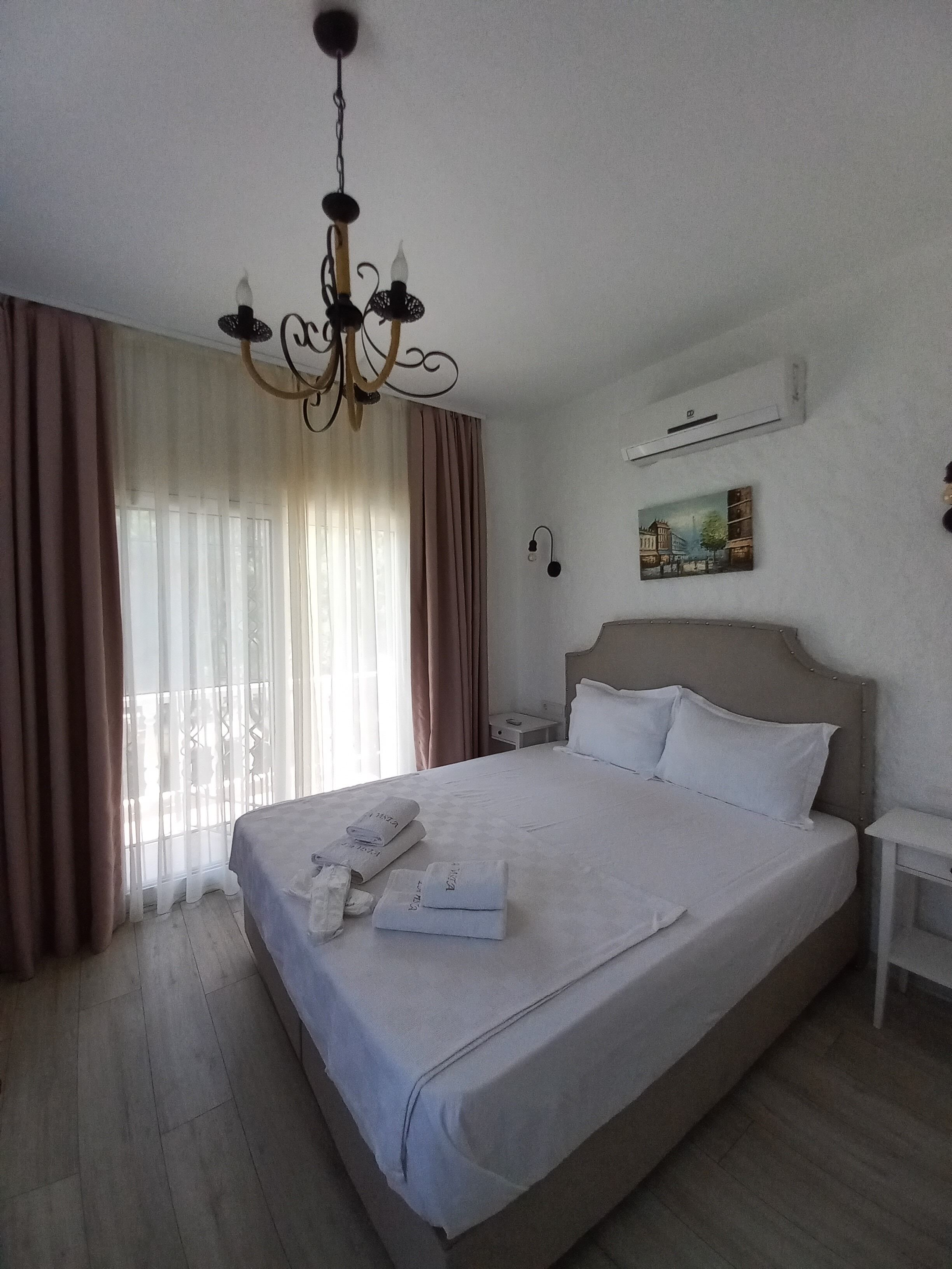 A hotel room with a large bed, a chandelier, a nightstand, a wall-mounted air conditioner, and a painting on the wall.