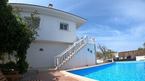 Un espace aménagé au bord de la piscine, avec une terrasse en bois, un bâtiment blanc doté d’un escalier et un ciel d’un bleu limpide.
