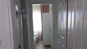 Un couloir étroit, dont le sol est recouvert de carreaux décoratifs, avec des portes blanches et une salle de bain visible, équipée de toilettes et d’un lit.