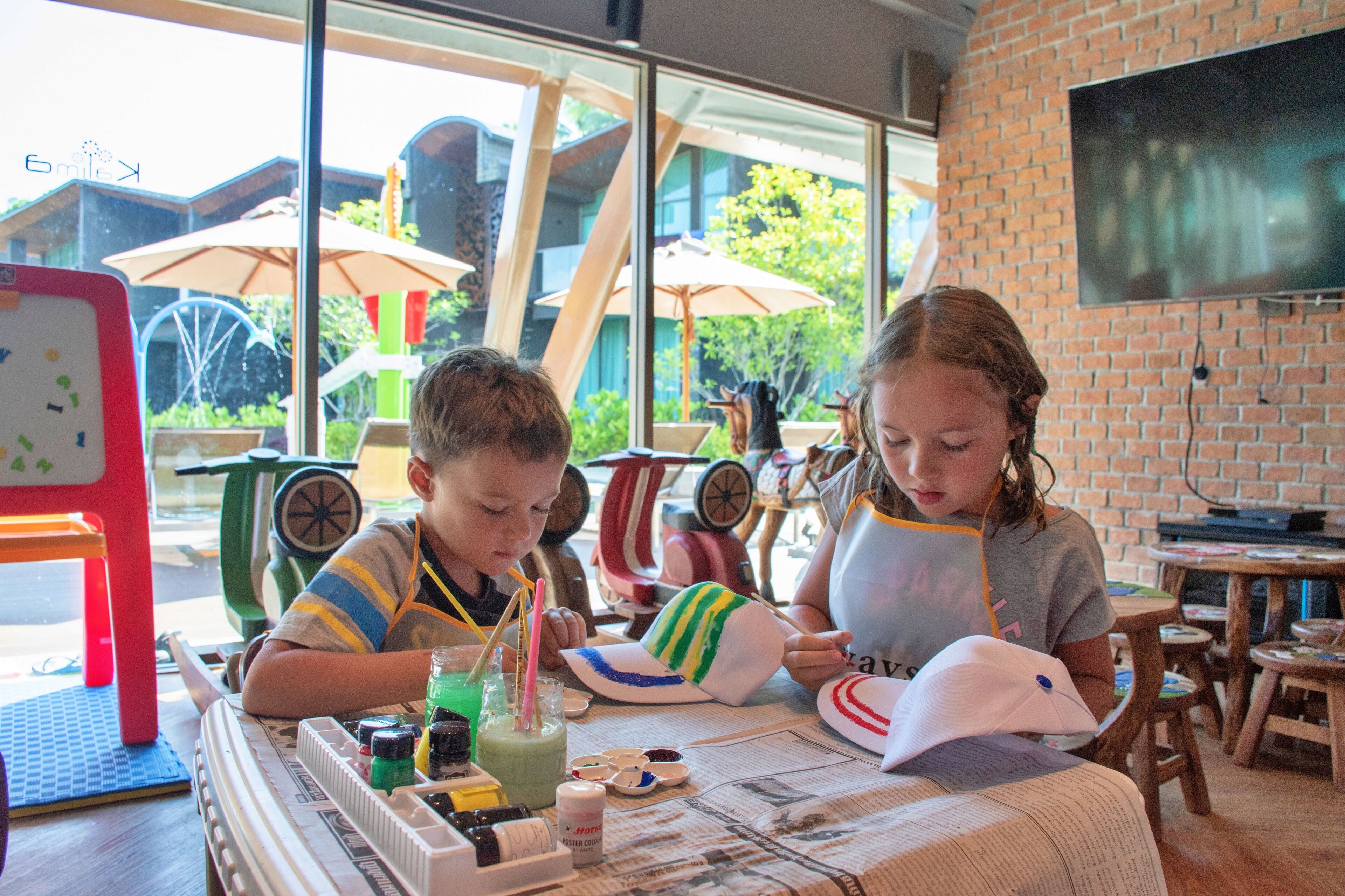 Two children at a table with art supplies, a TV on the wall, and outdoor umbrellas visible through the window.