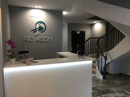 Un mostrador de recepción con un logotipo y el nombre 'RIO ASÓN' en la pared, una escalera y una orquídea en maceta.