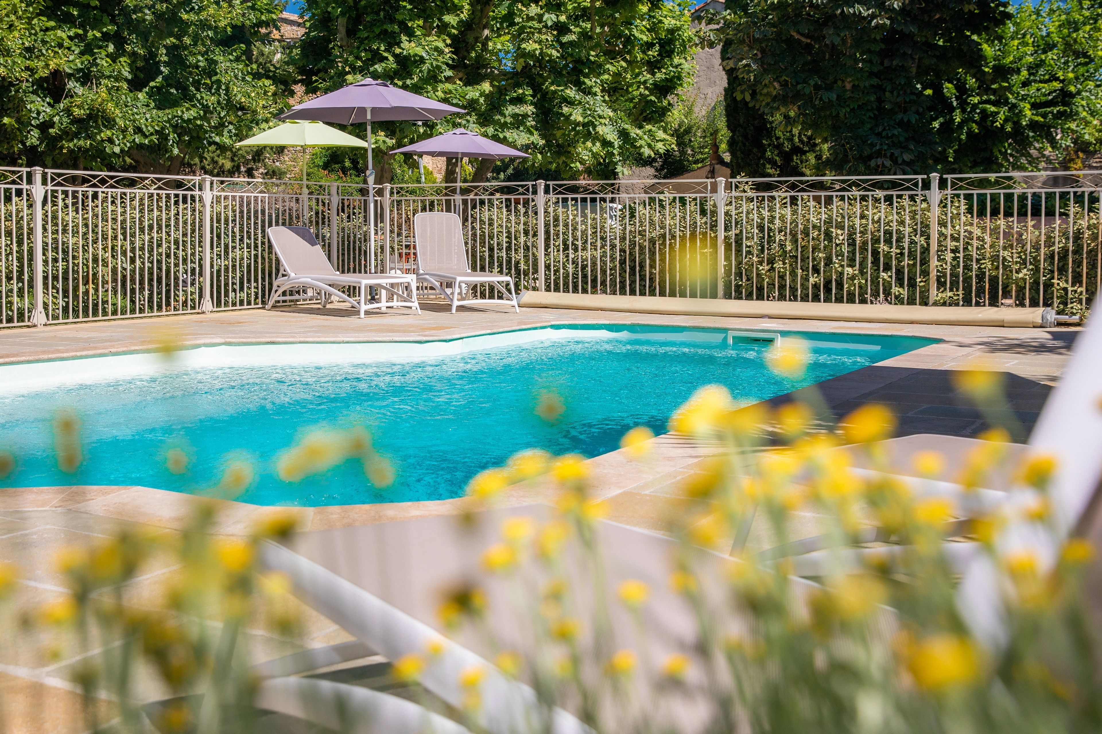 A swimming pool with two lounge chairs and umbrellas, surrounded by a fence and greenery.