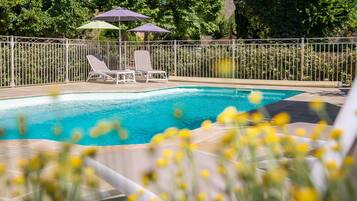 A swimming pool with two lounge chairs and umbrellas, surrounded by a fence and greenery.