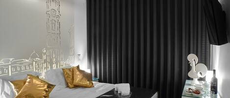 Une chambre d’hôtel moderne avec un lit en cuir noir, des coussins dorés, du linge de lit blanc et une table d’appoint en verre sur laquelle se trouvent une bouteille et des verres.
