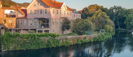 A large, historic building with a red-tiled roof, situated by a calm river.