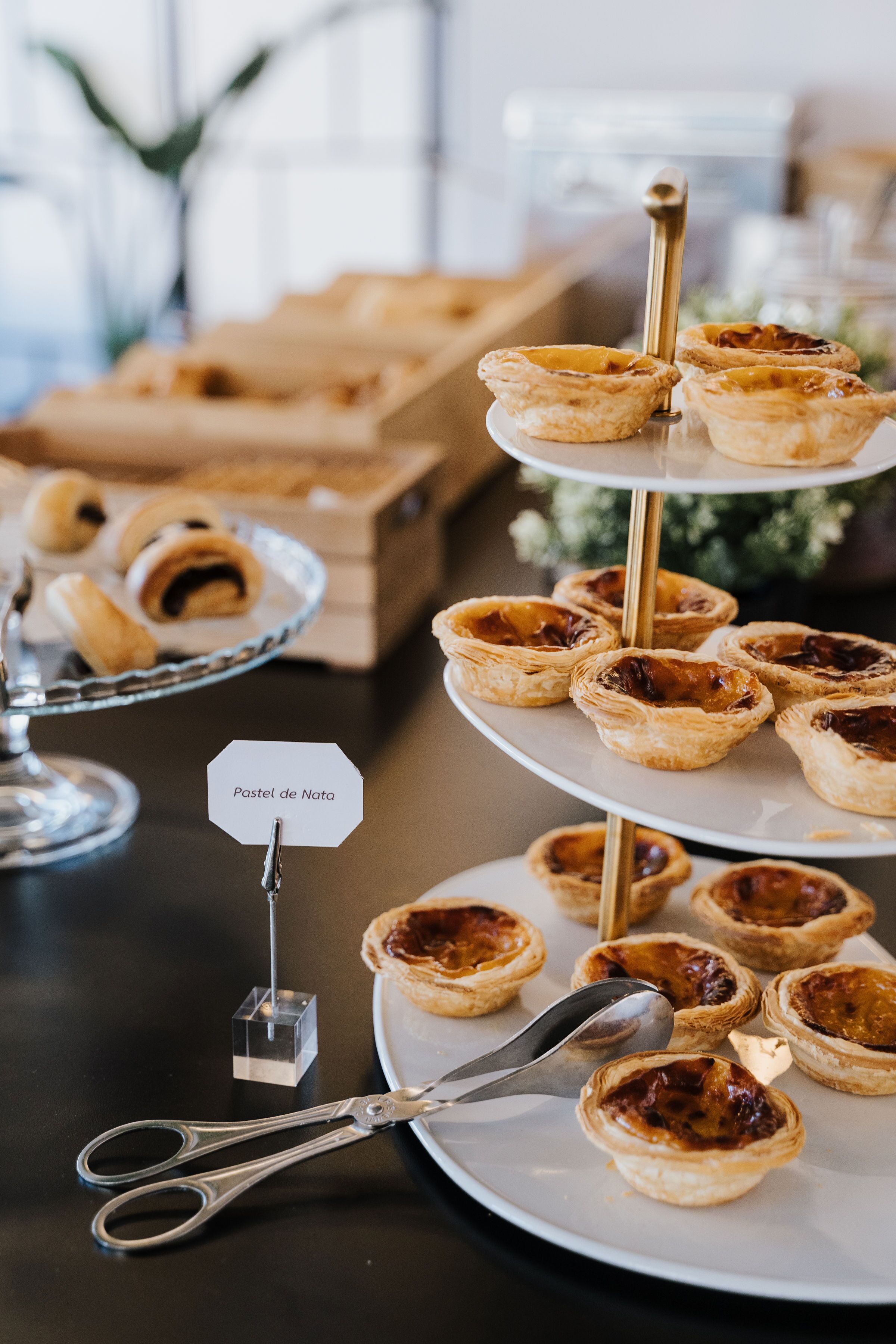 Un expositor escalonado con pasteles, incluyendo Pastel de Nata, sobre una mesa con un par de tijeras y una cuchara.