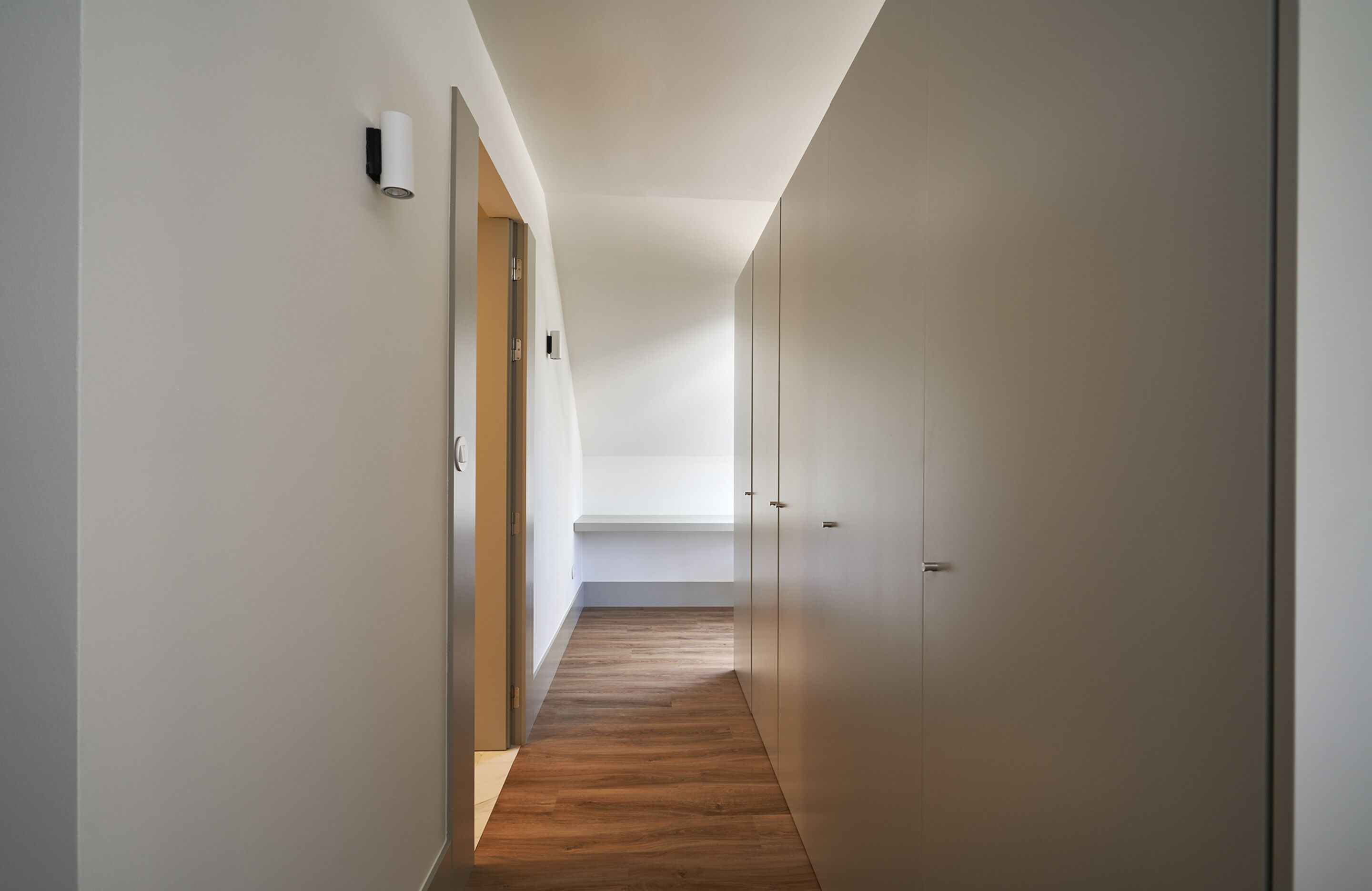 A narrow hallway with wooden flooring, white walls, and a door at the end.