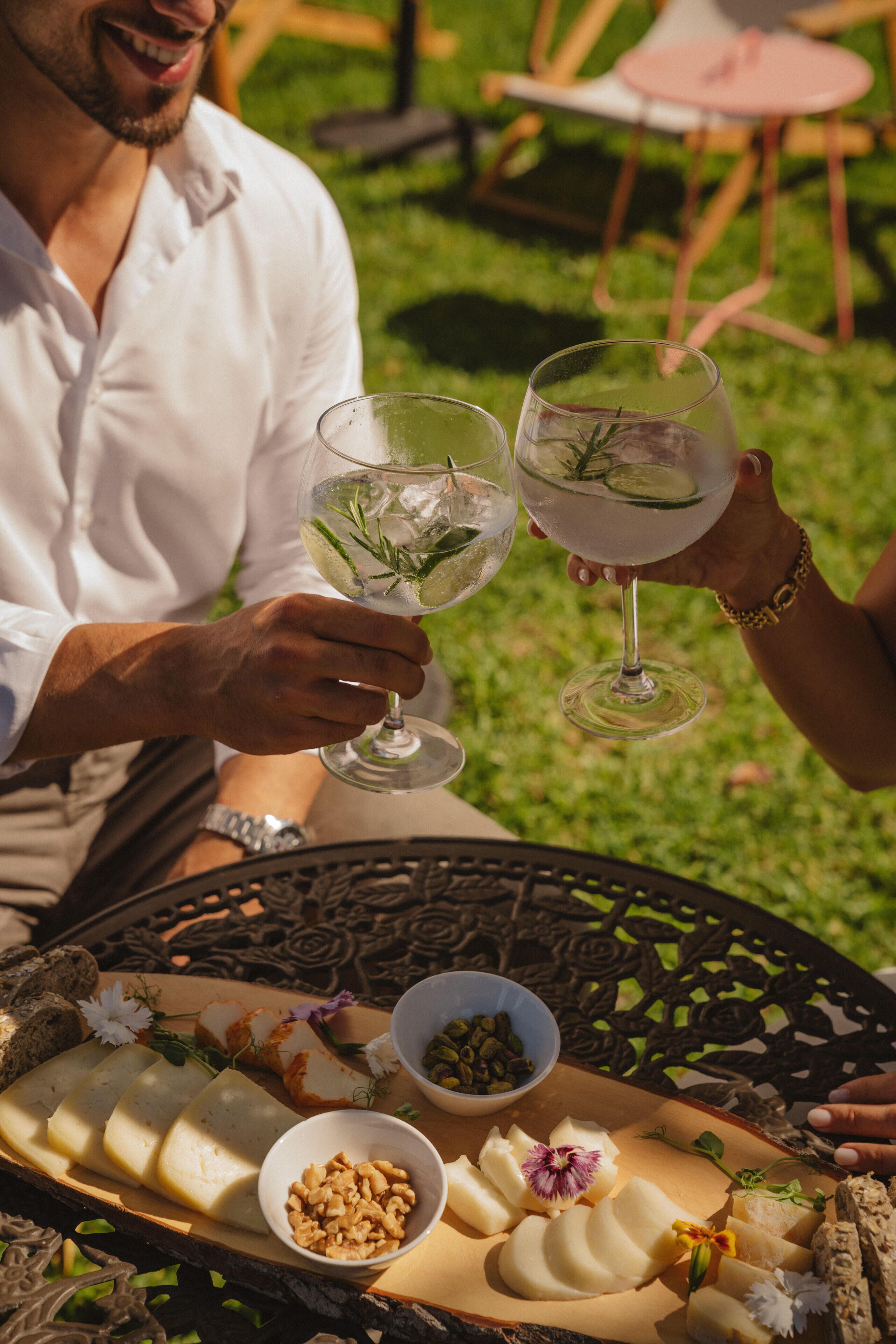 A person holding a glass of drink at an outdoor dining table with a variety of cheeses and nuts.