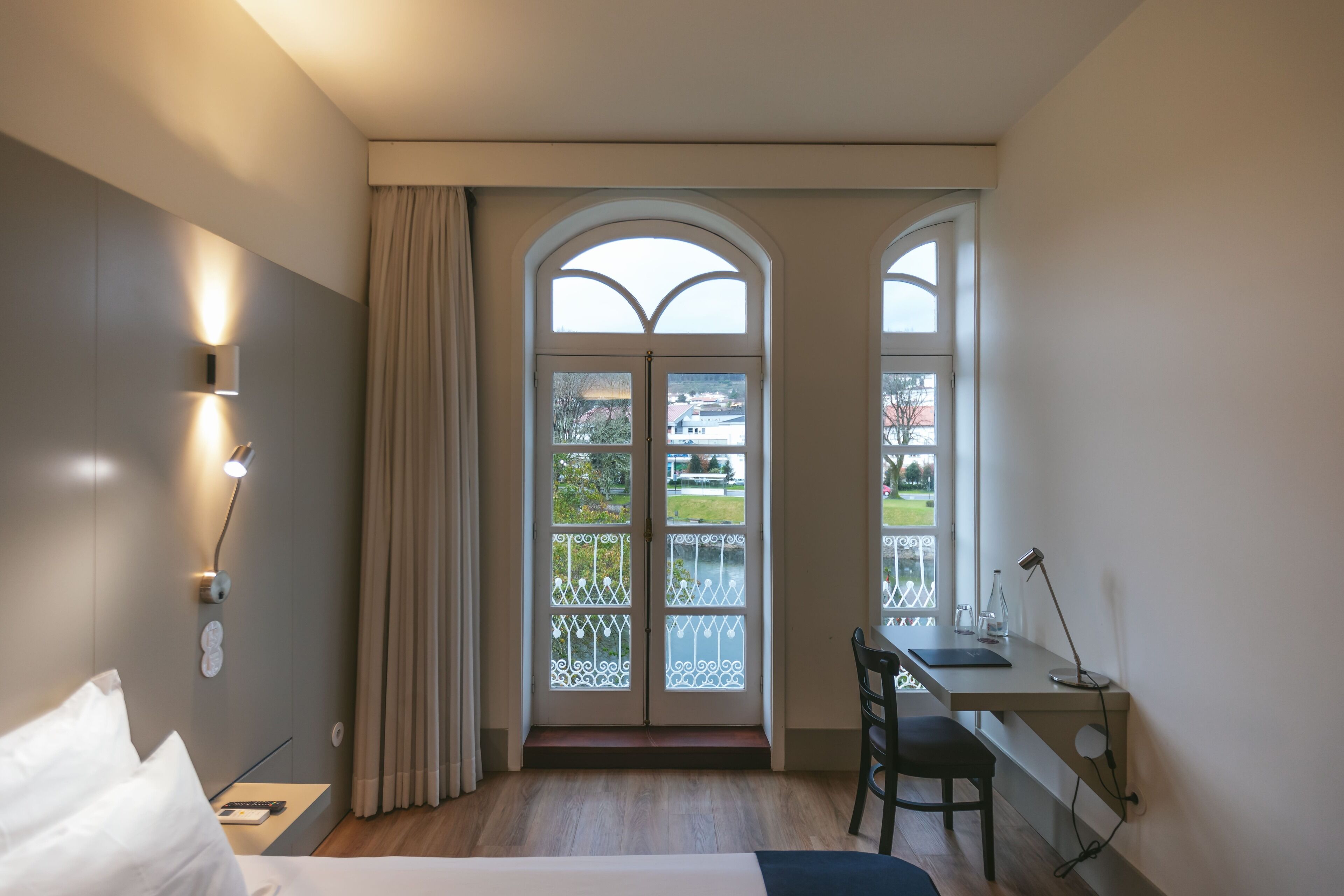 A hotel room with a bed, desk, and two windows with a view of a garden.