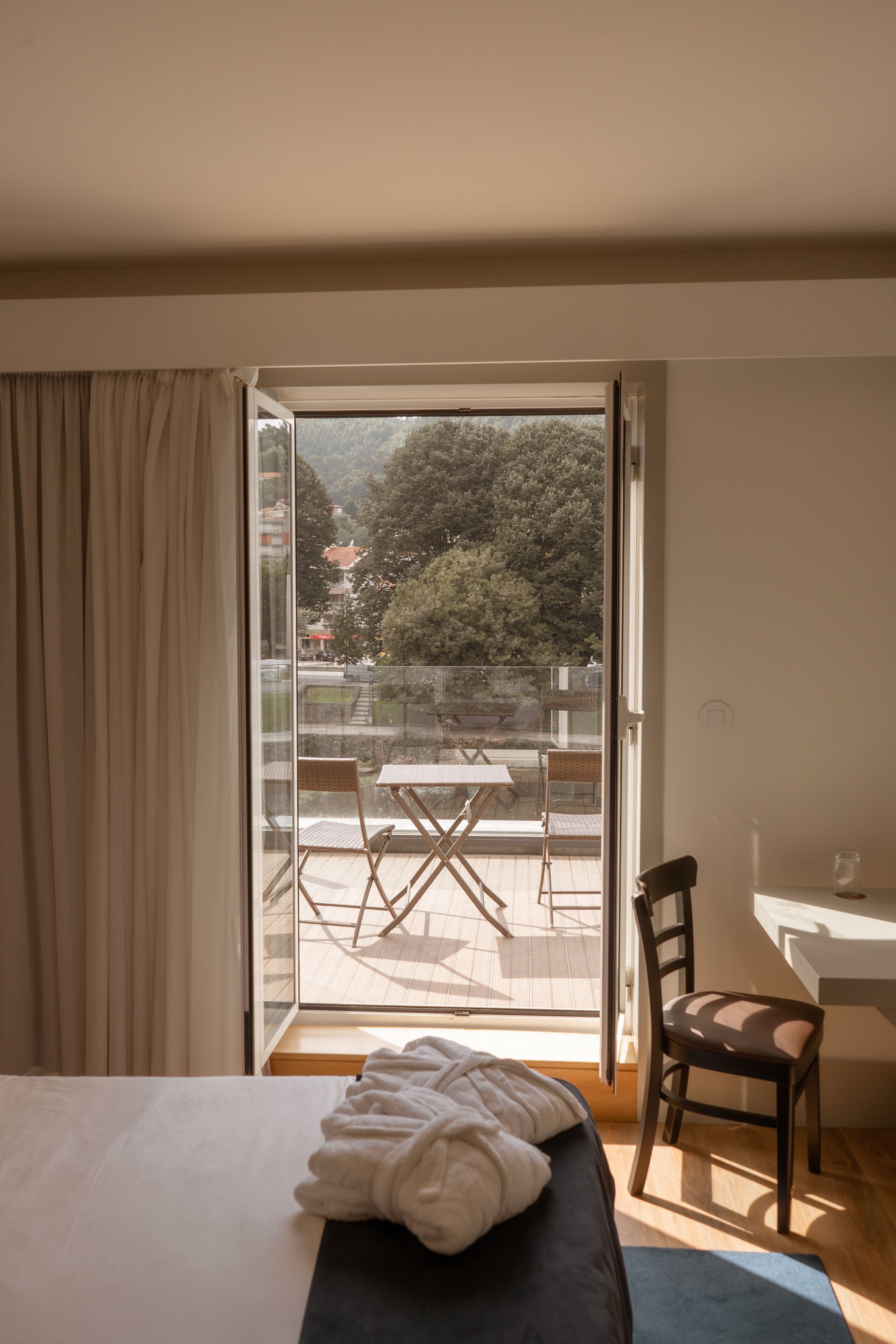Habitación de hotel con cama, una silla, una mesa y un balcón con vistas a árboles y edificios.