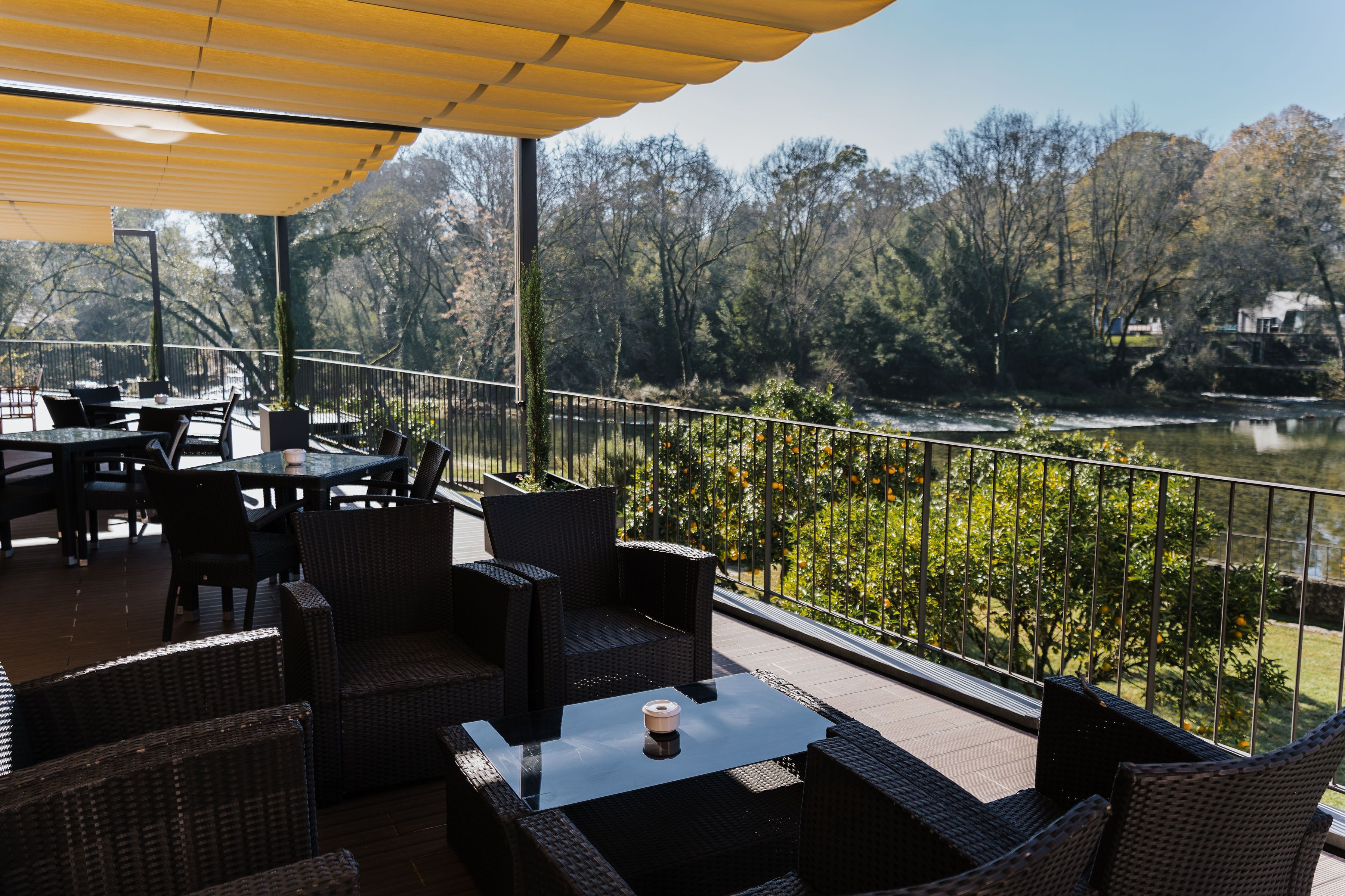 An outdoor patio with wicker furniture and a glass table, overlooking a river and trees.