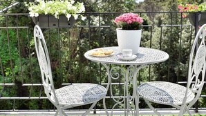 Une petite terrasse extérieure avec une table ronde et deux chaises, entourée d’une clôture en métal et de plantes en pot.