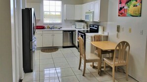 A compact kitchen with white cabinets, a refrigerator, a microwave, and a sink. There's a dining table with chairs and a painting on the wall. - Waters Edge Condo With Boat Slip (Freeport)