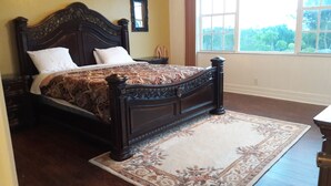 A bedroom with a dark wood bed, two white pillows, a patterned bedspread, a floral rug, a wooden nightstand, and a window with a view of greenery. - Waters Edge Condo With Boat Slip (Freeport)