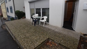 Un bâtiment à l’extérieur blanc, doté d’une porte-fenêtre coulissante et d’un petit espace extérieur aménagé avec deux chaises en plastique blanc et une table.