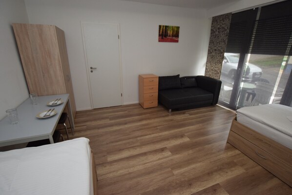 A room with a wooden floor, a bed, a sofa, a desk with a chair, and a window with a view of the outside. - AB Apartment 20 (Stuttgart)