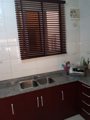 A kitchen with two stainless steel sinks, a granite countertop, dark wooden cabinets, a window with blinds, and a dish rack with utensils.