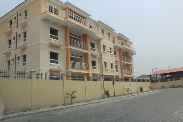 A multi-story residential building with balconies, a paved road in front, and a fence.