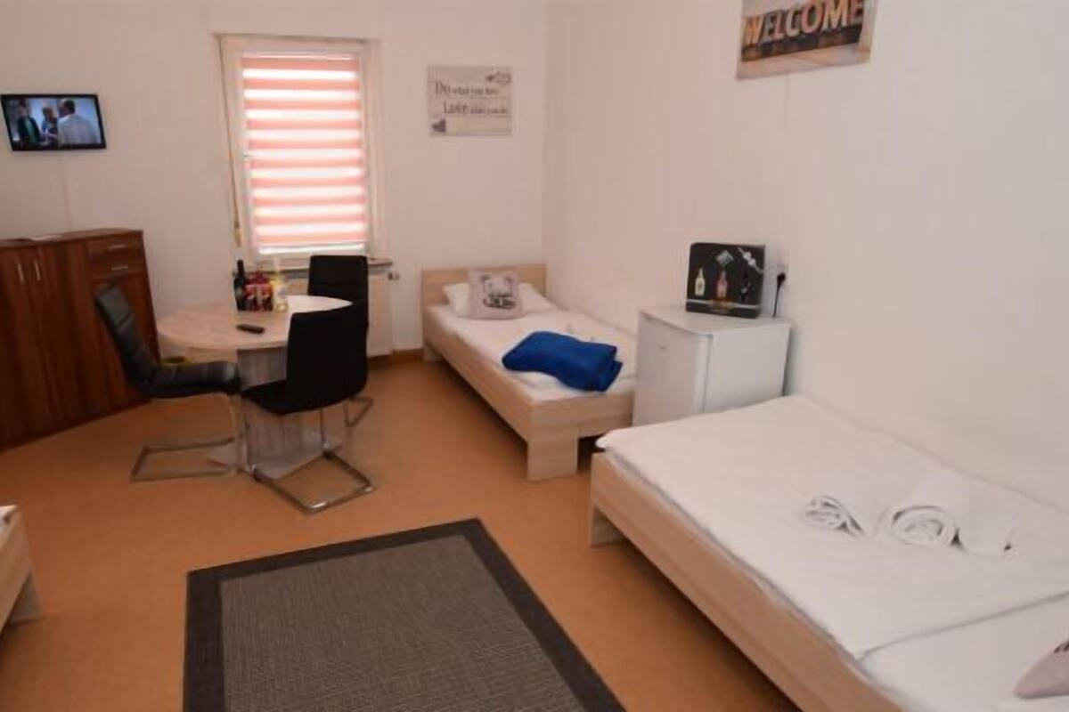 A compact room with a bed, a desk with a chair, a small table, and a TV on the wall.