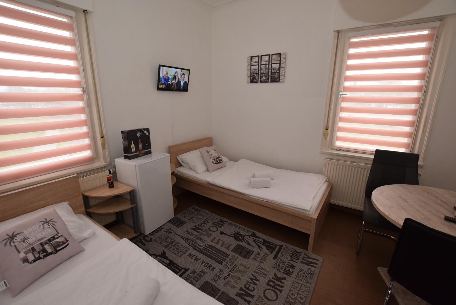 A compact hotel room with a single bed, a small desk, a chair, a TV mounted on the wall, and a window with blinds.