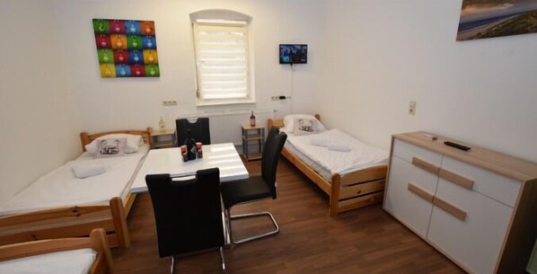 Una habitación tipo dormitorio con dos camas individuales, un escritorio, una silla, un armario pequeño y una ventana con persianas.