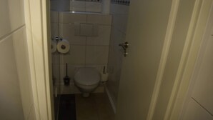Une petite salle de bain étroite, équipée de toilettes, d’un rouleau de papier toilette et d’un sèche-mains.