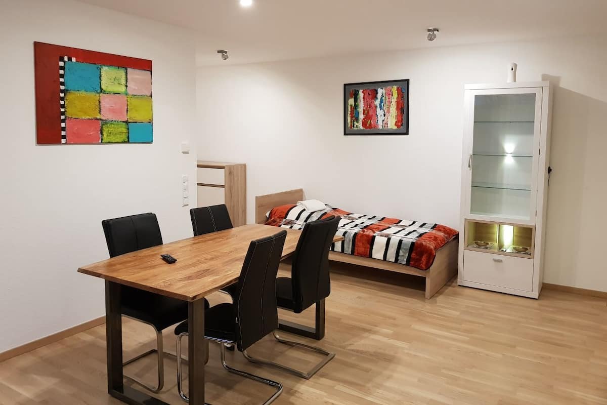 Une pièce comprenant une table à manger en bois, des chaises noires, un lit recouvert d’une couverture à rayures, une vitrine et deux tableaux abstraits accrochés aux murs.