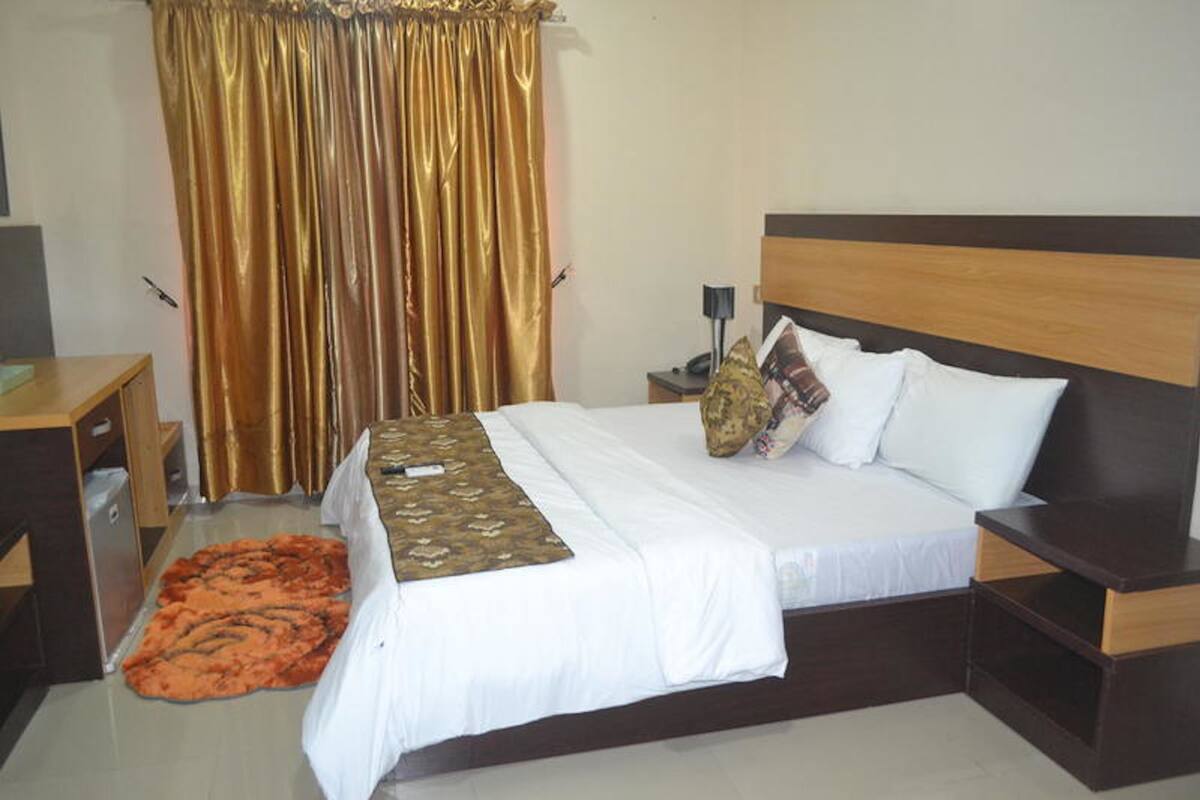 A hotel room with a bed, a nightstand, a lamp, a wooden headboard, and a curtain.