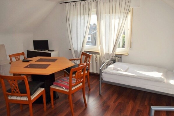 A small, cozy room with a wooden dining table, four chairs, a single bed, a TV, and a window with curtains.