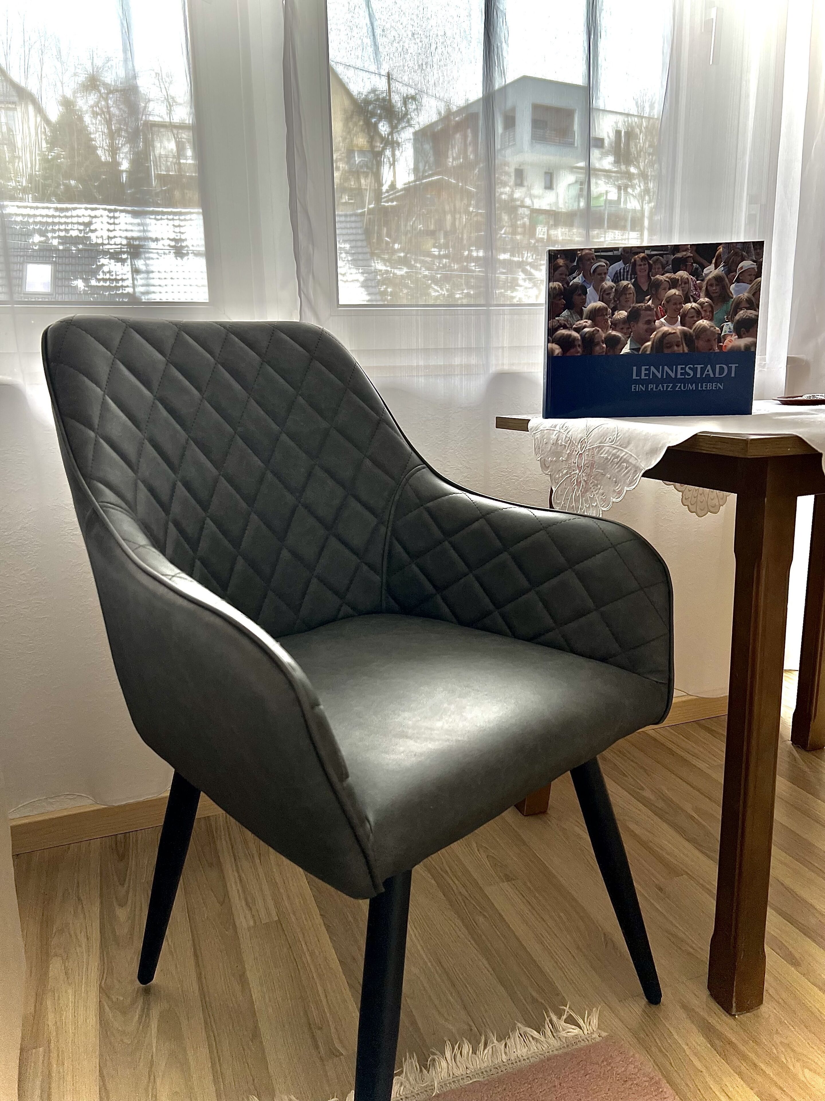 Un fauteuil moderne doté d’un dossier matelassé et de pieds en bois, placé dans une pièce avec de grandes fenêtres et une affiche accrochée au mur.