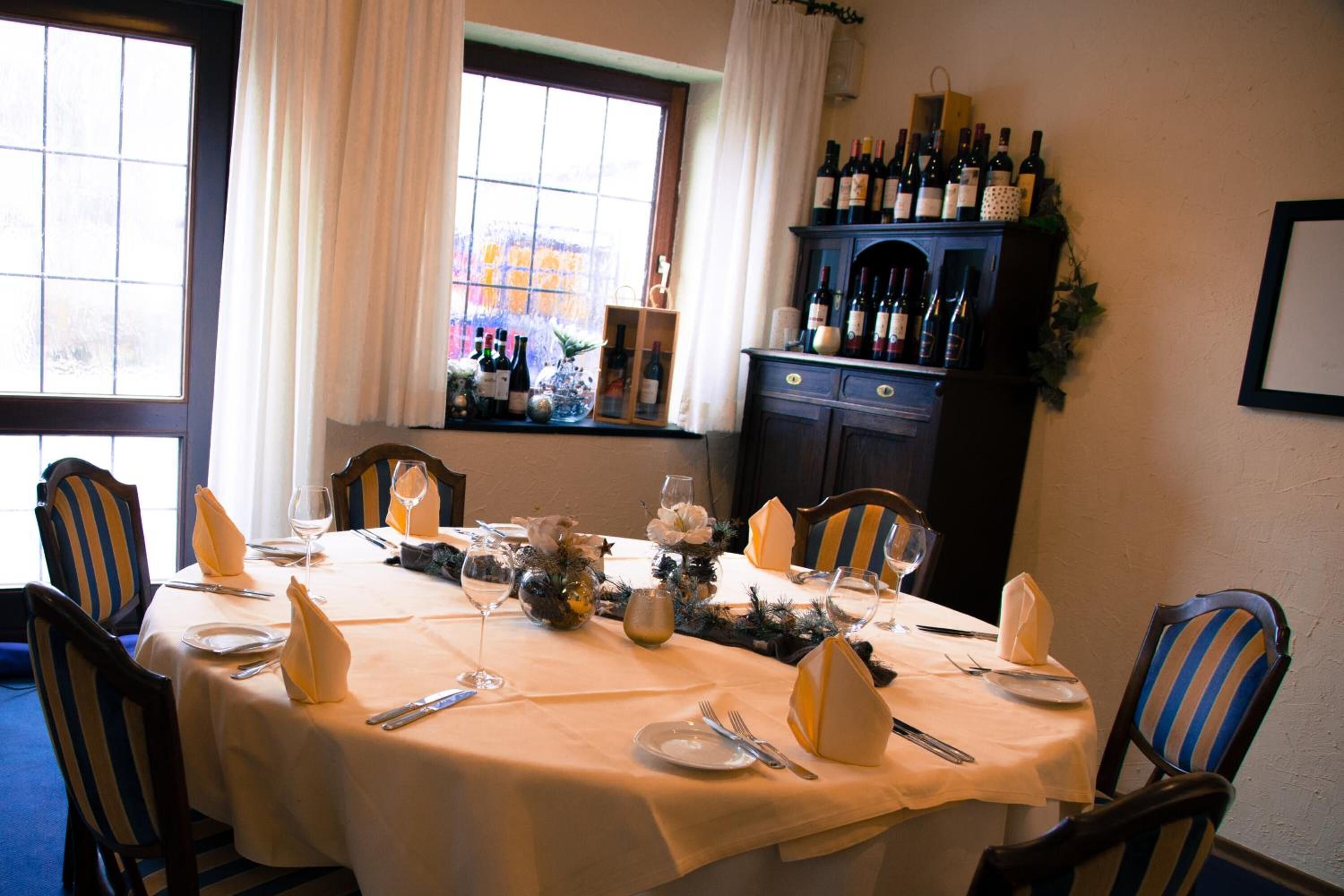 A dining table set for six with a white tablecloth, wine bottles on a shelf, and a window with curtains.