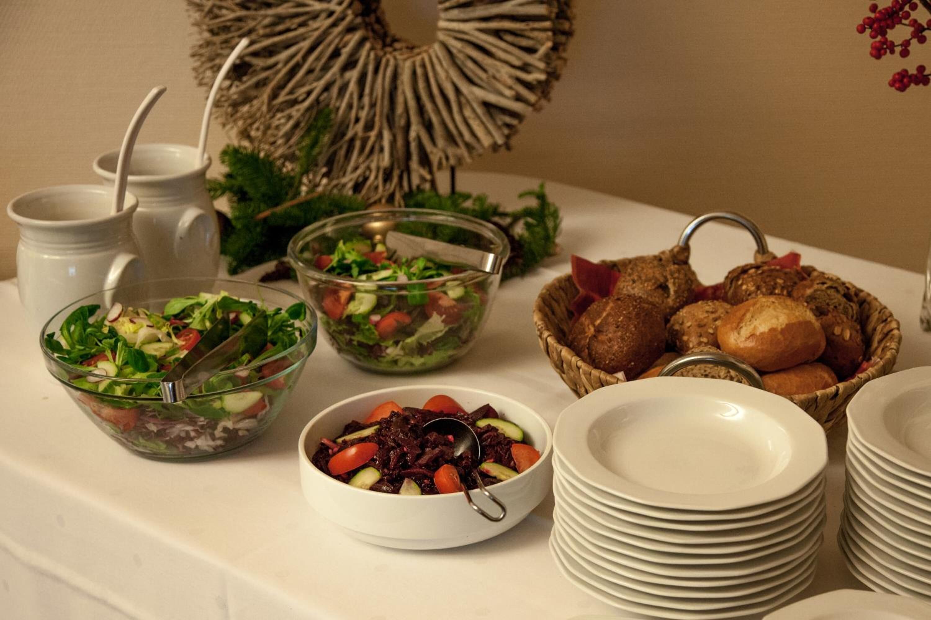 Une table de buffet proposant divers plats, notamment des salades et du pain, ainsi que des piles d’assiettes blanches.