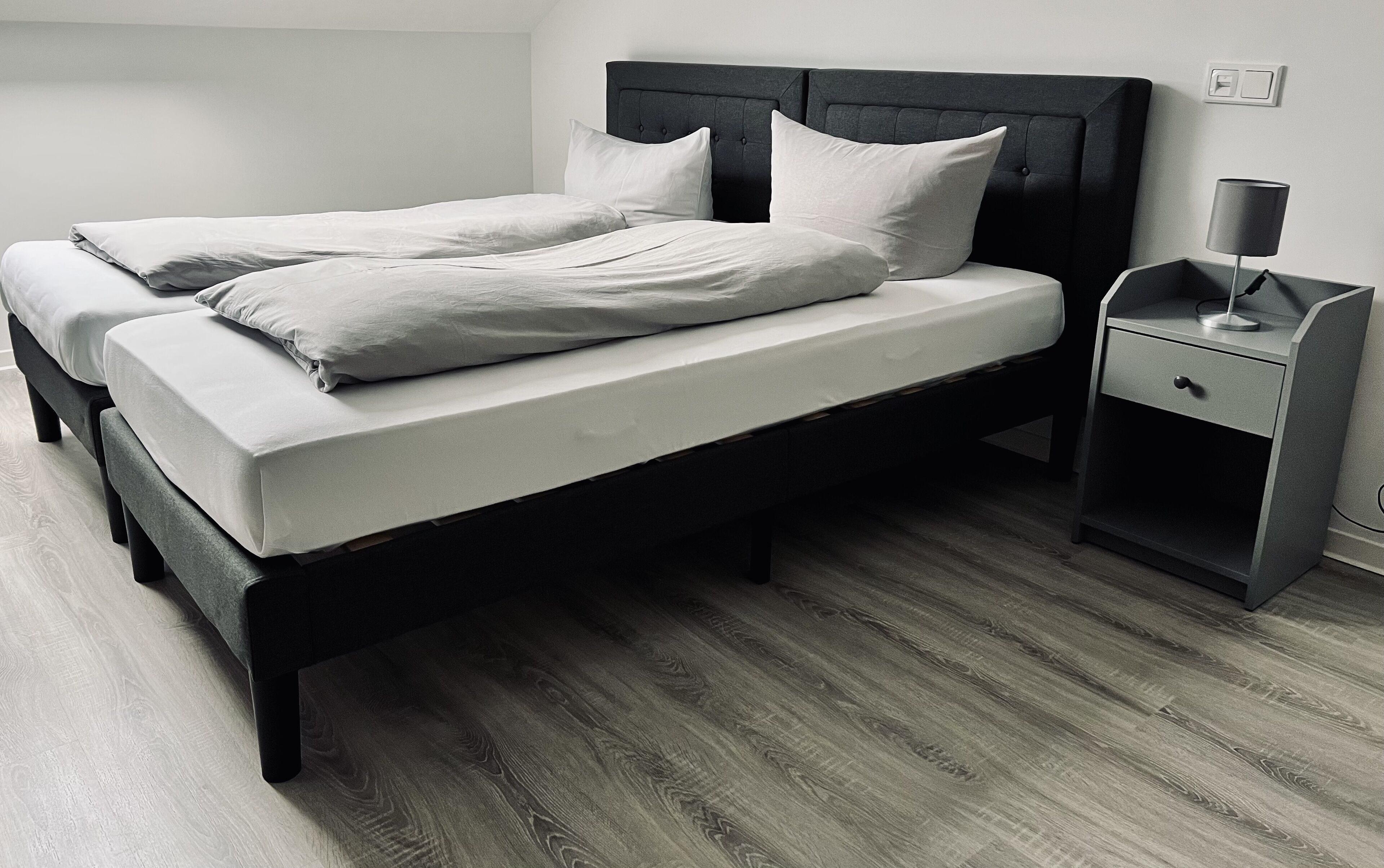 A double bed with a dark headboard and white bedding, a bedside table with a lamp, and a wooden floor.