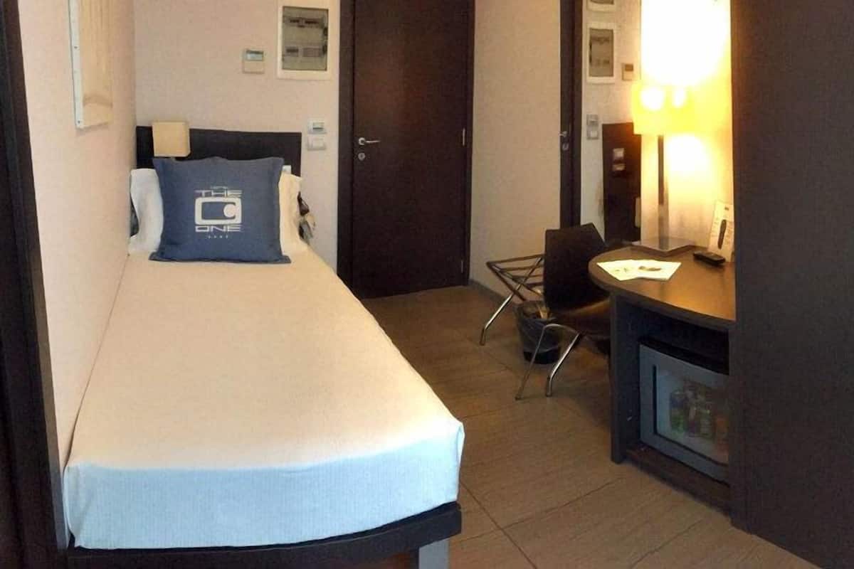 A hotel room with a single bed, a desk with a chair, a small fridge, and a mirror.