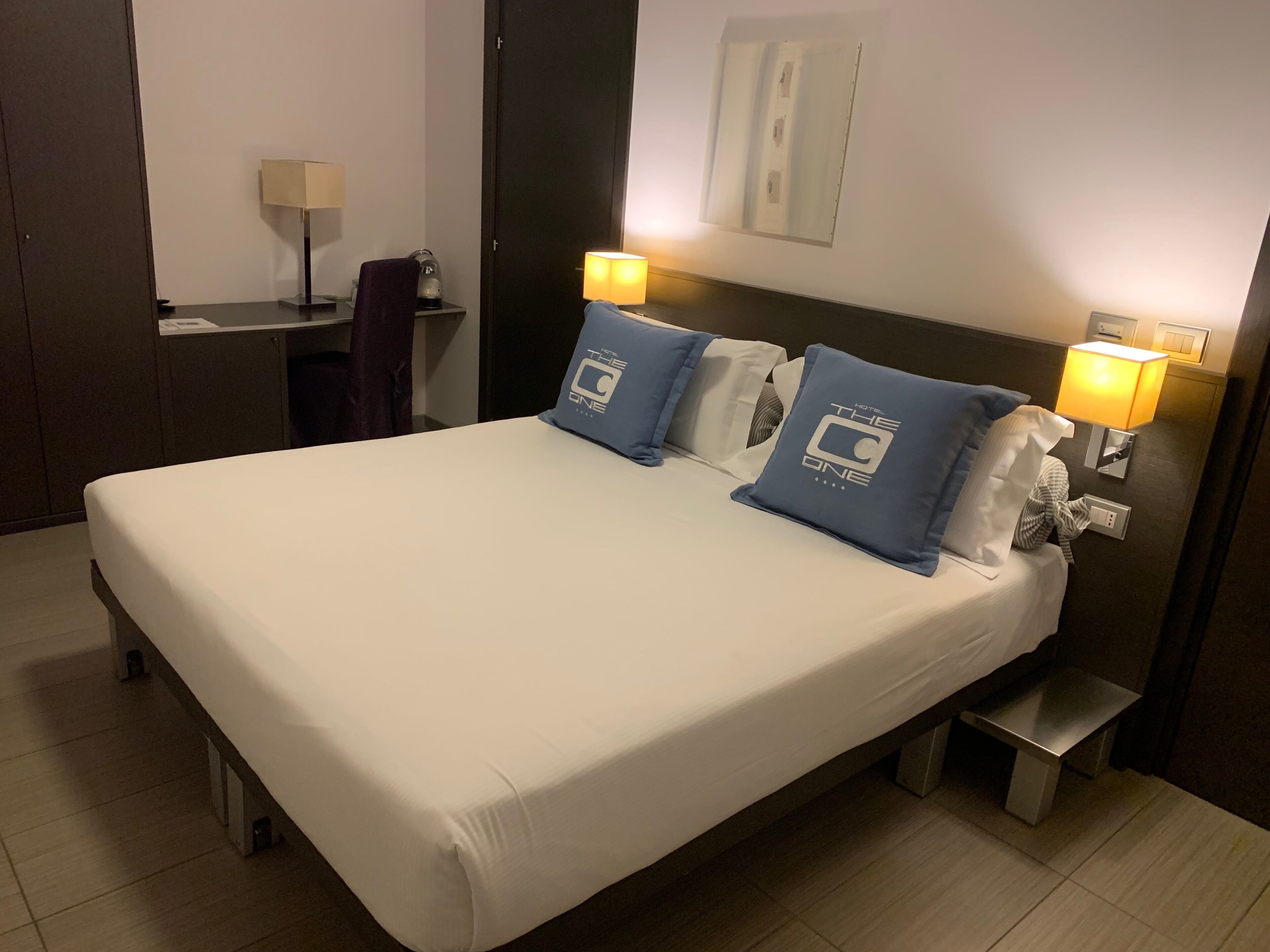 Una habitación de hotel con una cama grande, dos almohadas con el logotipo del hotel, lámparas de noche, una mesita pequeña y una mesita de noche.