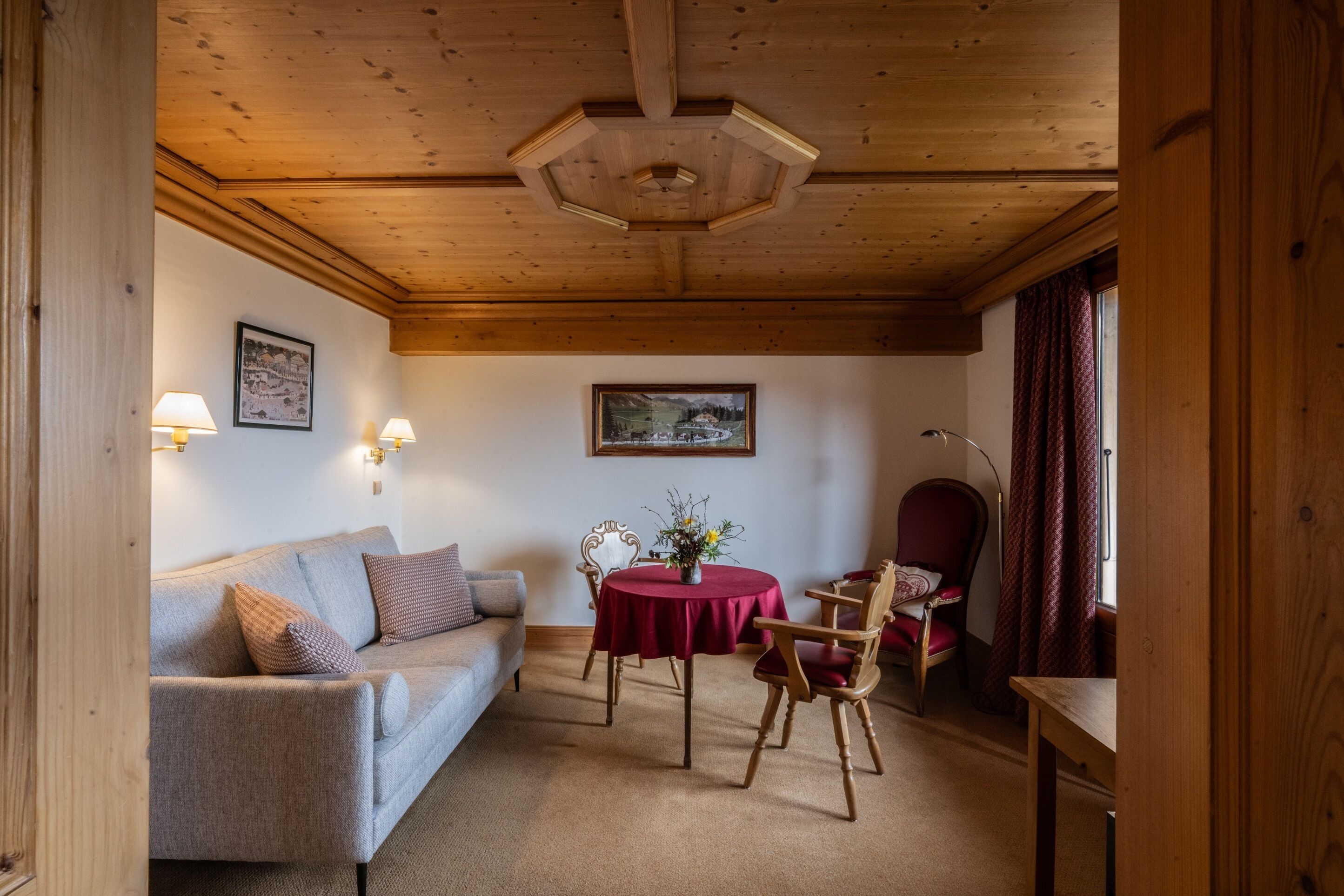 A cozy room with a wooden ceiling, a sofa, a dining table with chairs, and a chair with a red cushion.