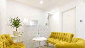 A room with a tufted yellow sofa, a small white table with a bottle and glasses, and a potted plant. - Best Location Rooms (Split)