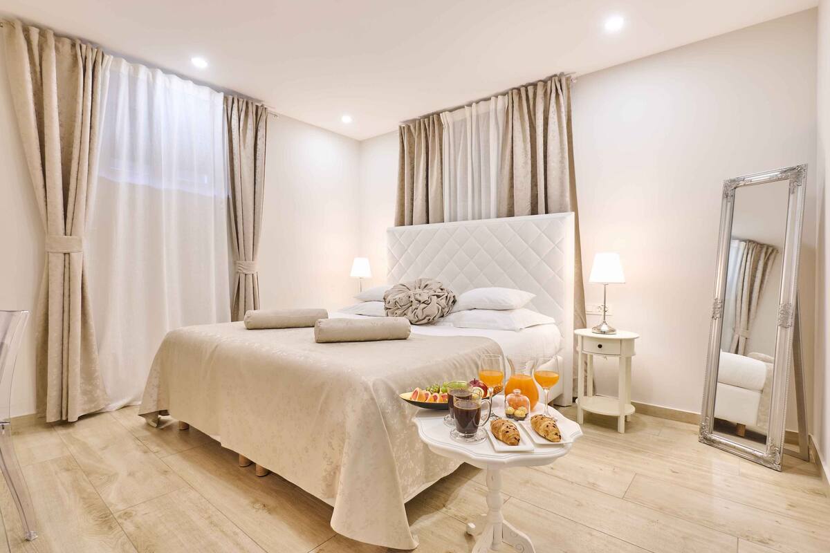 A neatly made bed with a quilted headboard, a bedside table with a lamp, and a tray of breakfast items.